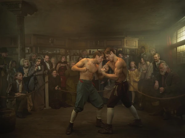 Two shirtless male boxers face off in an indoor ring surrounded by a crowd in vintage clothing. The scene is dimly lit, creating a dramatic effect.
