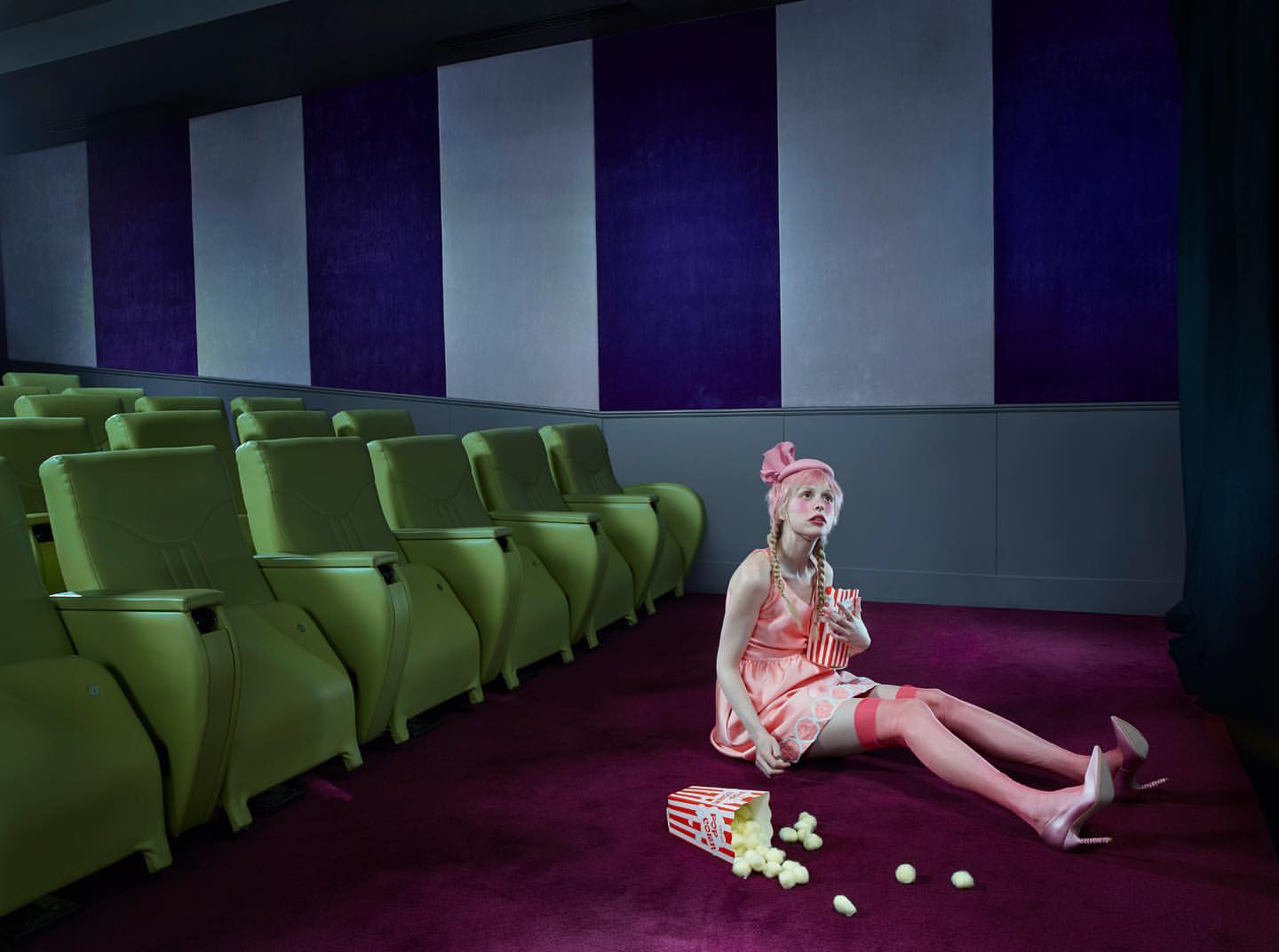 Woman in a pink dress sits cross-legged on a purple carpet at the movie theater, with scattered popcorn and green theater chairs.