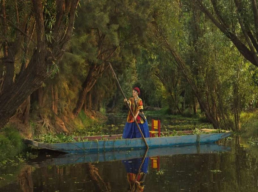 Woman in traditional dress rows a wooden boat through a lush, green canal, surrounded by trees reflecting in the water.