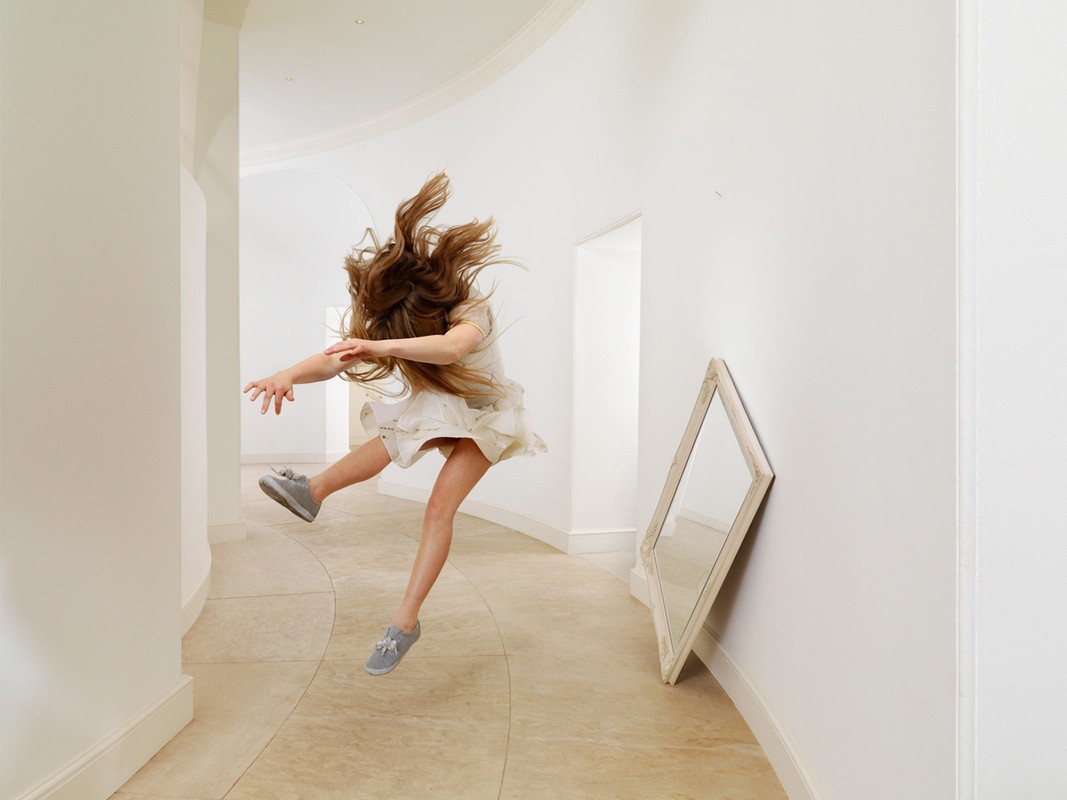 A young girl caught mid-air mid-jump in a bright white hallway with a leaning mirror by Julia Fullerton-batten.