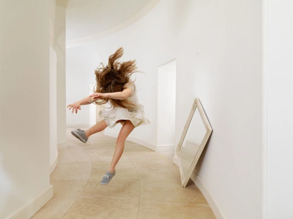 A young girl caught mid-air mid-jump in a bright white hallway with a leaning mirror by Julia Fullerton-batten.