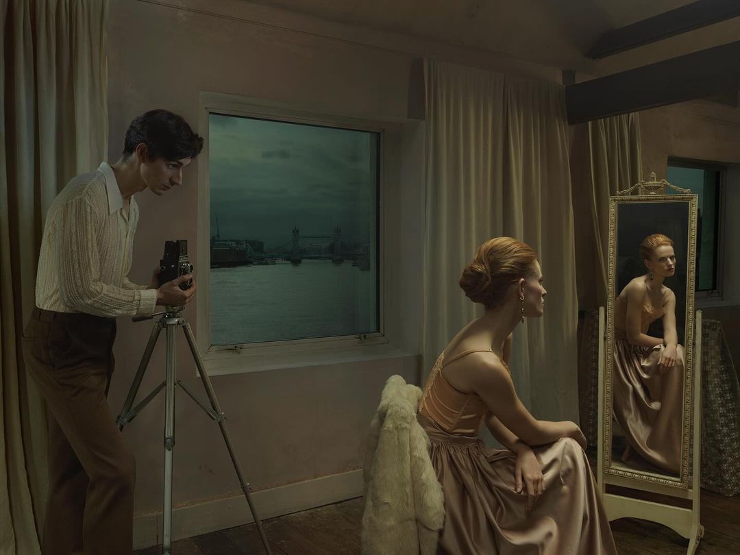 Indoor scene features a man photographing a woman near a window with a cityscape view and a mirror reflecting the woman seated.