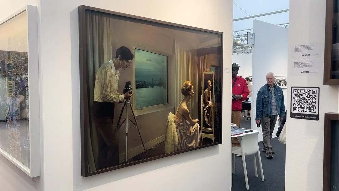 Framed photograph at an art fair shows a man photographing a woman looking in a mirror, with a window view of a city skyline.