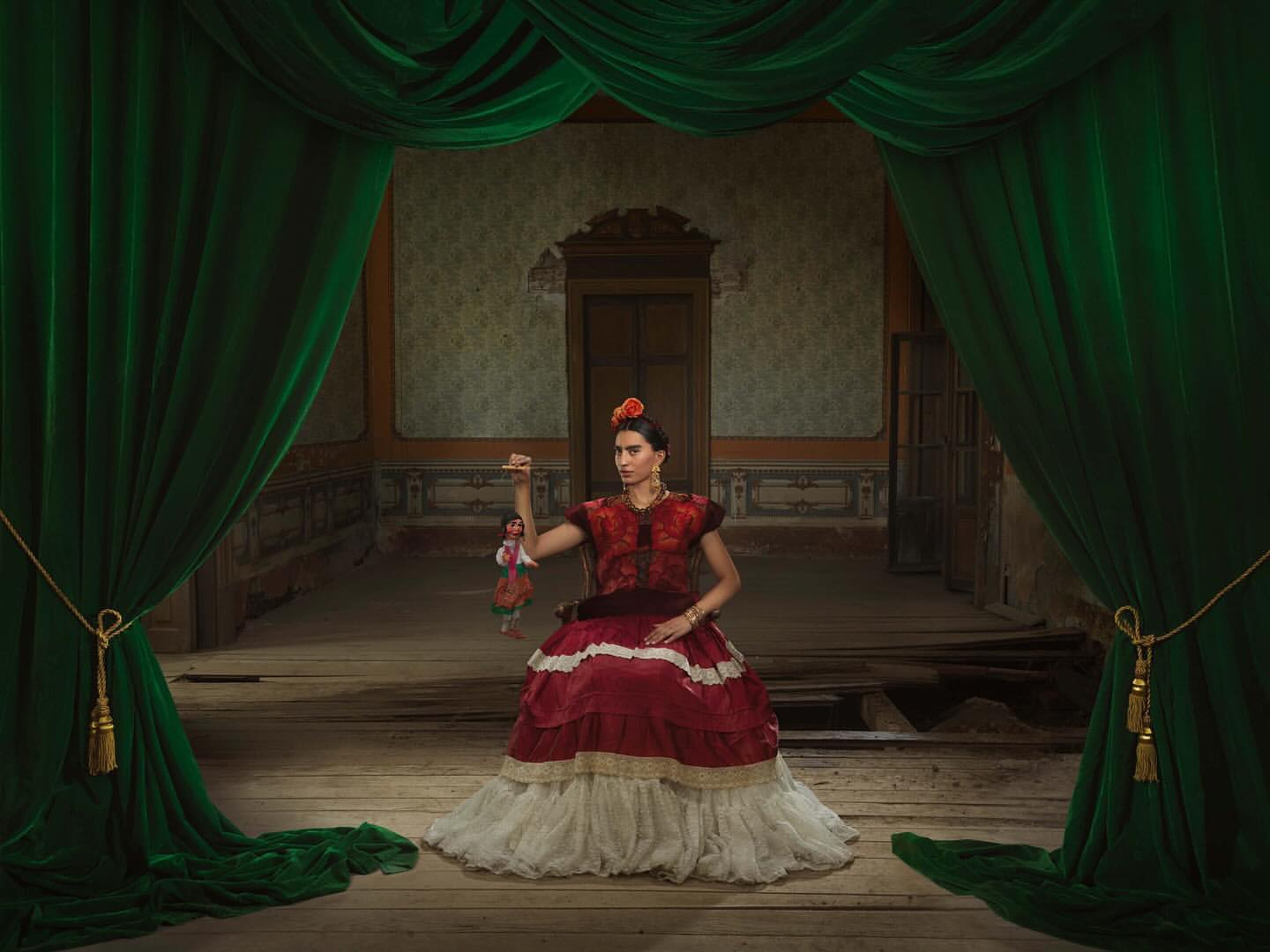A woman in a red and white dress poses with a doll on a stage with green curtains, reminiscent of a Frida Kahlo portrait.