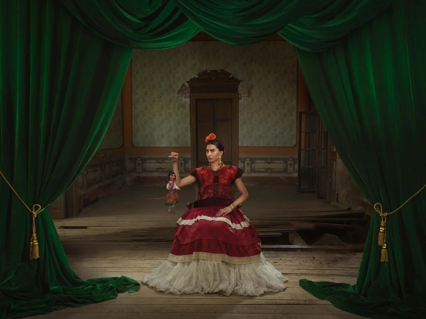 A woman in a red and white dress poses with a doll on a stage with green curtains, reminiscent of a Frida Kahlo portrait.
