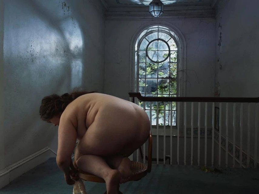 Nude adult woman poses on a small stool in a dimly lit decaying home with a large arched window and overgrown foliage outside.