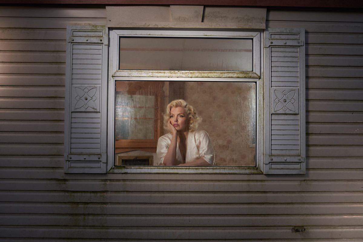 Woman with blonde hair and pale skin looks pensively through a window, leaning on her hand, in an indoor setting with daylight.