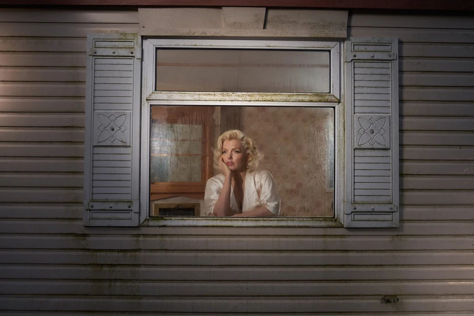 Woman with blonde hair and pale skin looks pensively through a window, leaning on her hand, in an indoor setting with daylight.