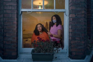 Two women by a window of a brick building, one in an orange shirt and one in a pink shirt wearing a striped mask, photographed by Julia Fullerton-Batten.