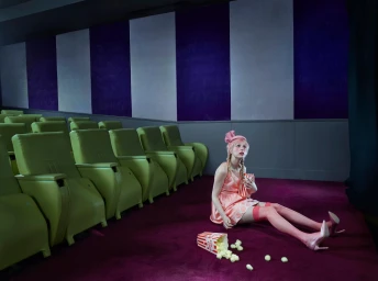 A woman in a pink dress sitting on a purple cinema floor beside spilled popcorn, facing rows of green seats.