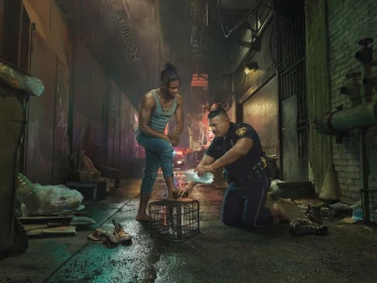 A police officer kneels in a gritty alley to wash the foot of a barefoot man standing with one foot on a crate, photographed by Julia Fullerton-Batten.
