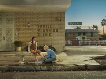 A woman kneeling washes another seated woman foot outside a clinic building with a motel sign visible in the background.