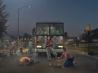 Group of people standing by a bus on a suburban street at dusk with a woman kneeling to wash another woman's foot in a basin