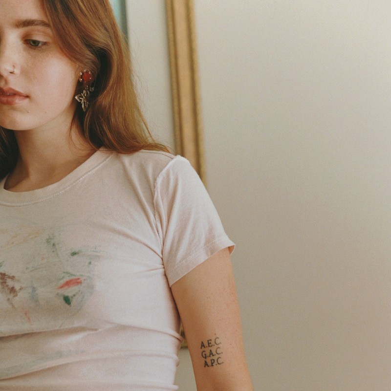 Close-up portrait of a young woman with blonde hair wearing a white t-shirt featuring a subtle print and small lettering tattoo on her arm