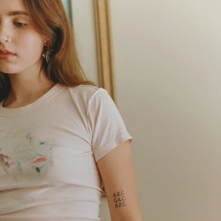 Close-up portrait of a young woman with blonde hair wearing a white t-shirt featuring a subtle print and small lettering tattoo on her arm