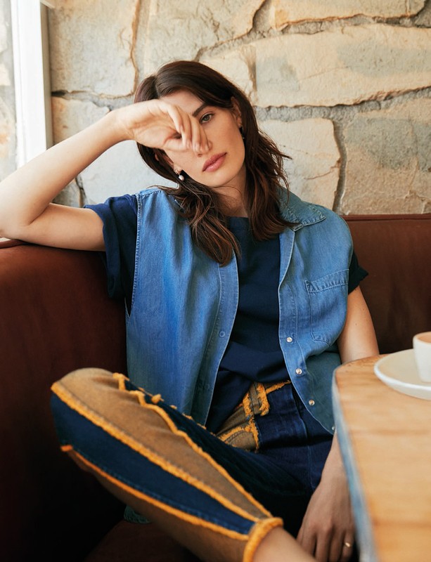 Brunette woman in casual denim vest poses indoors with stone wall background, seated at table with coffee, in soft, natural light.