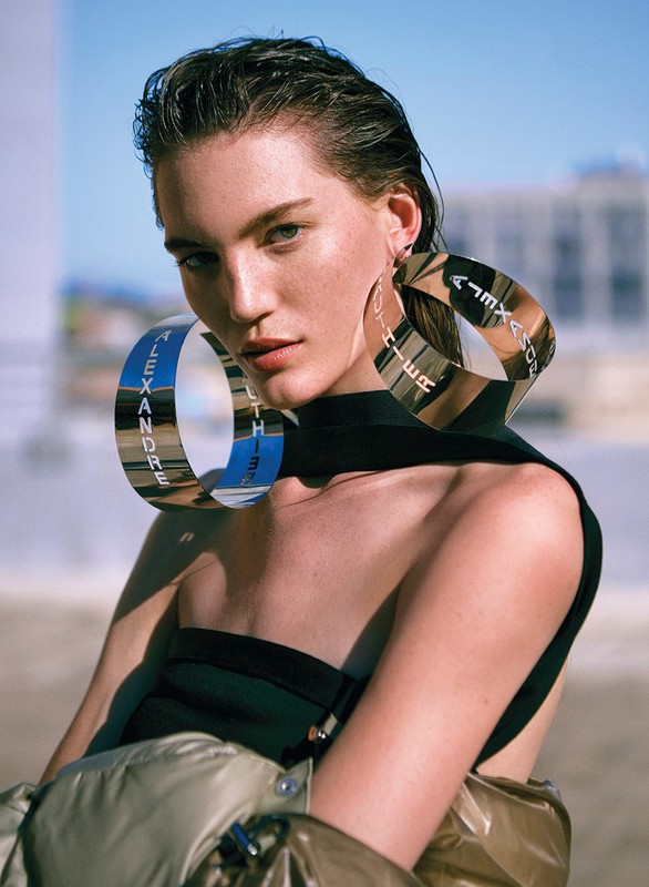 Fashion portrait of a woman with wet hair wearing hoop earrings that have lettering; she also wears a black top and gold jacket.