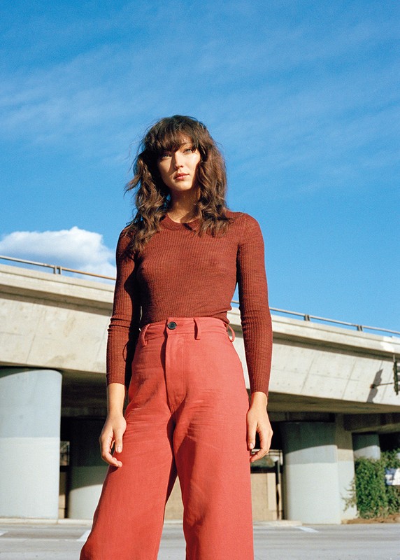 Woman in long-sleeved top and orange wide-leg pants stands outside with a blue sky background and an elevated bridge overhead.