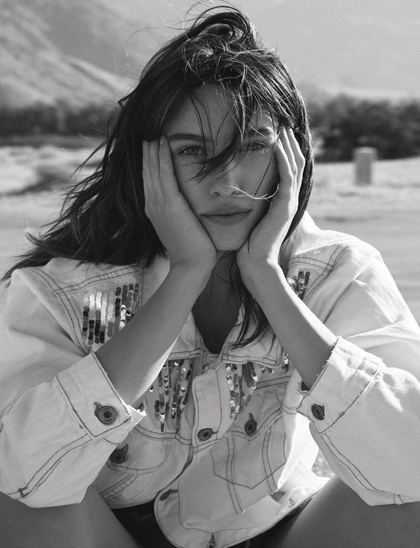 Monochrome portrait of a young woman with long hair posing outdoors, wearing a denim jacket with sequins and her hands on her face.