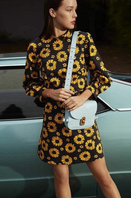 A stylish woman in a black and yellow floral dress poses next to a blue car, accessorized with a light blue purse.