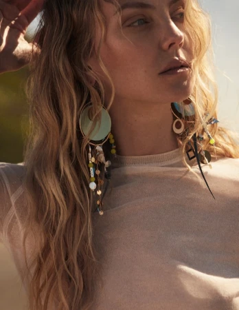 Close-up portrait of a woman with blonde hair wearing large statement earrings and a cream top under natural sunlight.