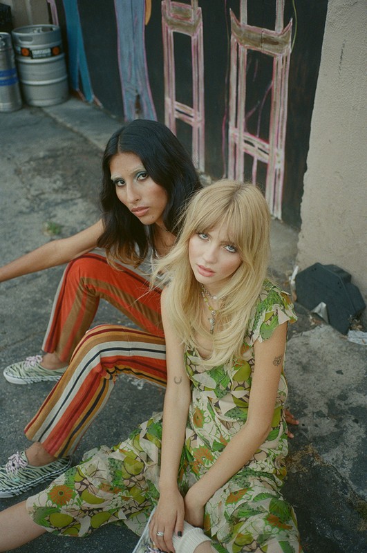 Two women sit outdoors in stylish, floral-patterned and striped clothing, posing against a textured wall with painted artwork.