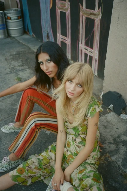 Two women sit outdoors in stylish, floral-patterned and striped clothing, posing against a textured wall with painted artwork.