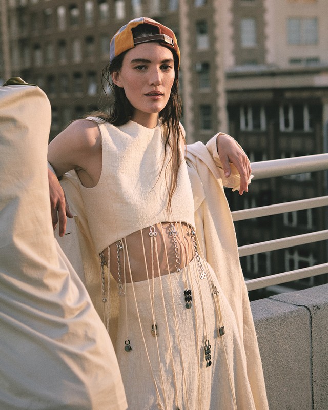 Two women wearing matching off-white outfits pose on a rooftop with city buildings in the background; focus on the woman in frame right.