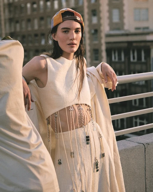 Two women wearing matching off-white outfits pose on a rooftop with city buildings in the background; focus on the woman in frame right.