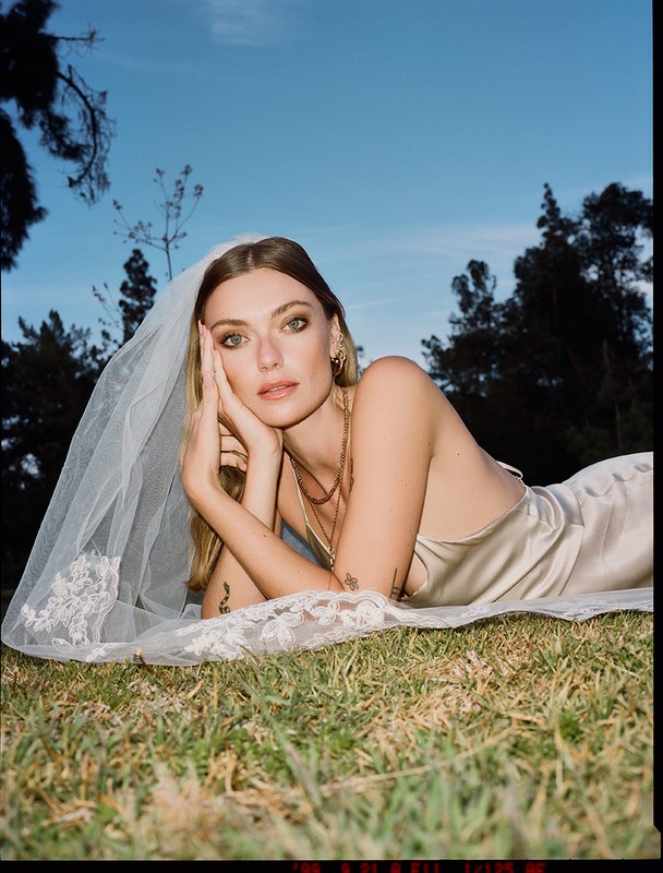 A woman in a wedding dress and veil laying in a grassy field, looking at the camera under a blue sky framed by trees.
