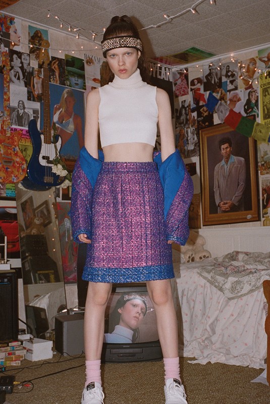 Young woman in white crop top and purple mini skirt poses in retro bedroom with posters and vintage TV set. Fashion editorial shot.