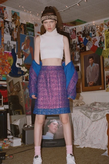 Young woman in white crop top and purple mini skirt poses in retro bedroom with posters and vintage TV set. Fashion editorial shot.