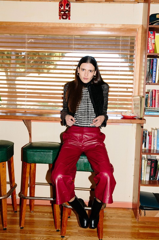 A young woman sits on a barstool in front of a window wearing a black-and-white top, red leather pants, and black boots in an indoor setting.