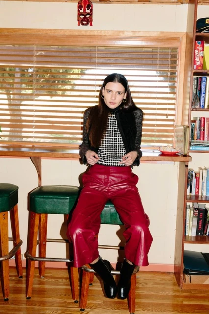 A young woman sits on a barstool in front of a window wearing a black-and-white top, red leather pants, and black boots in an indoor setting.