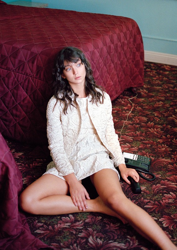 A young woman in a white dress and jacket sits on a floral carpet in front of a burgundy bed, holding a vintage telephone.