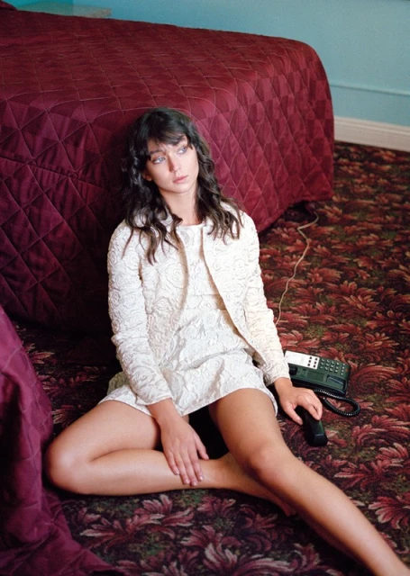 A young woman in a white dress and jacket sits on a floral carpet in front of a burgundy bed, holding a vintage telephone.