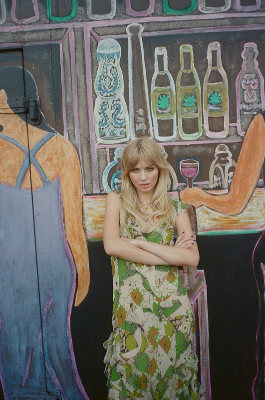 Young woman in floral dress stands with arms crossed against a backdrop of painted bottles in an outdoor setting, medium shot.