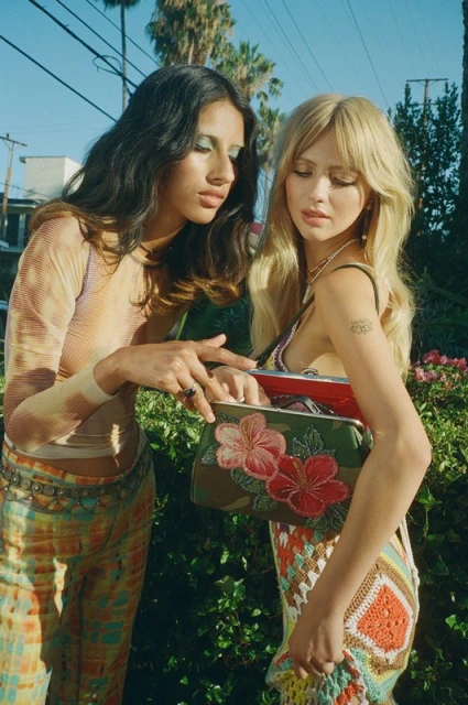 Two women pose outdoors, showcasing embroidered floral purses in a close-up shot with palm trees and a blue sky.