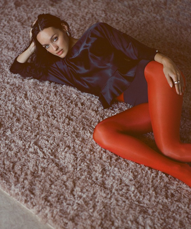Brunette woman lying on a shag carpet wearing a black satin blouse and red tights, indoors in a studio setting, looking at the camera.