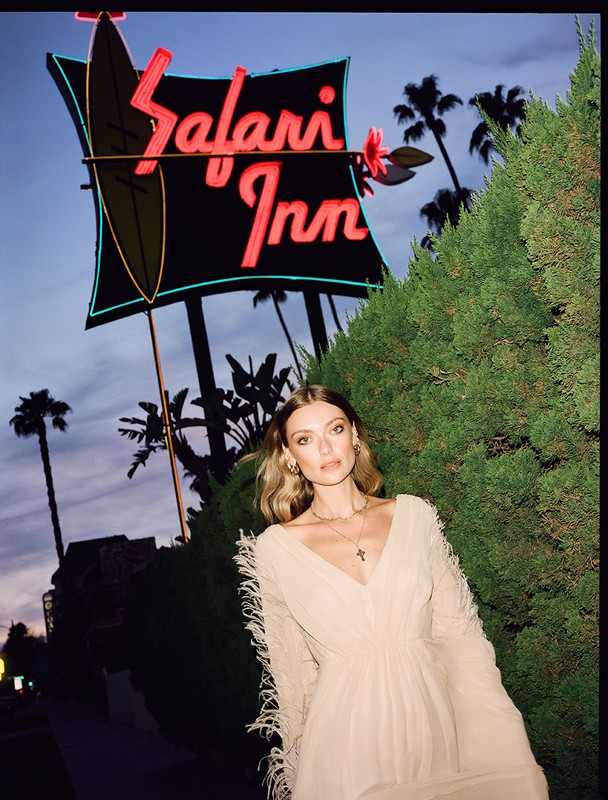 A stylish woman in a long dress stands in front of the Safari Inn sign at dusk, framed by lush greenery and silhouetted palm trees.