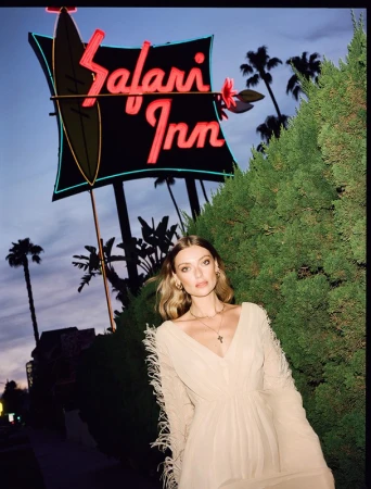 A stylish woman in a long dress stands in front of the Safari Inn sign at dusk, framed by lush greenery and silhouetted palm trees.