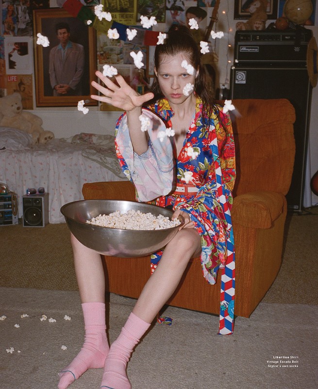 A young woman in a colorful shirt sits in an orange chair, throwing popcorn in the air, with various objects in the background.