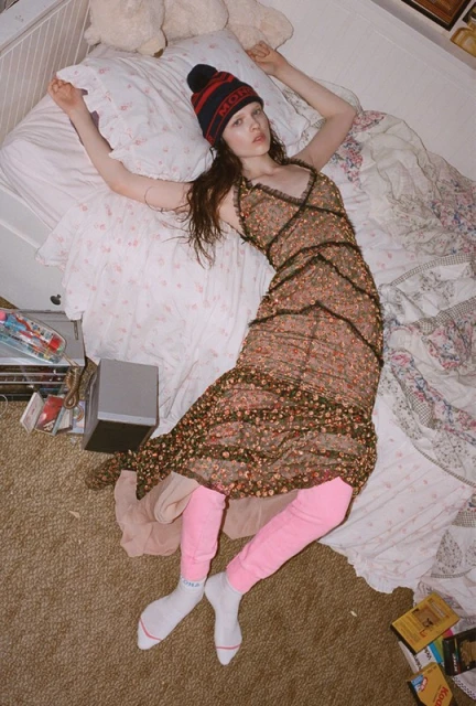 Overhead view of a young woman lying on a bed in a floral dress, red knit hat, and pink leggings, in a bedroom with a brown carpet.