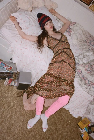 Overhead view of a young woman lying on a bed in a floral dress, red knit hat, and pink leggings, in a bedroom with a brown carpet.