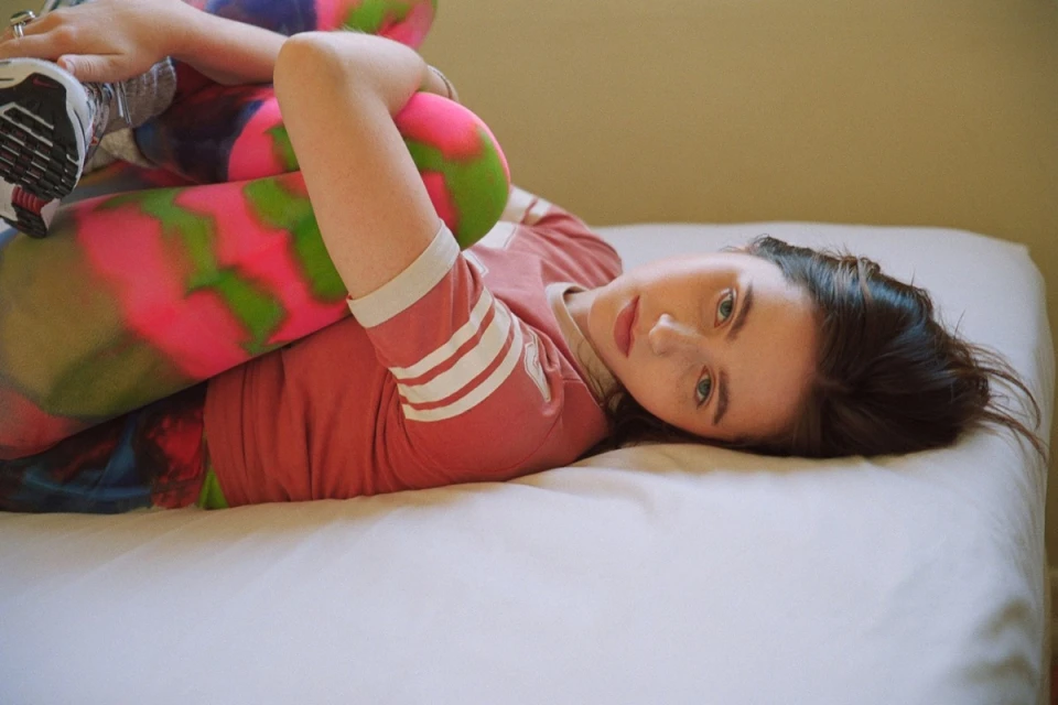 Woman in activewear lies on white bed, knees to chest, smiling at camera. Colorful leggings, striped sleeves, indoor setting. Fitness lifestyle.