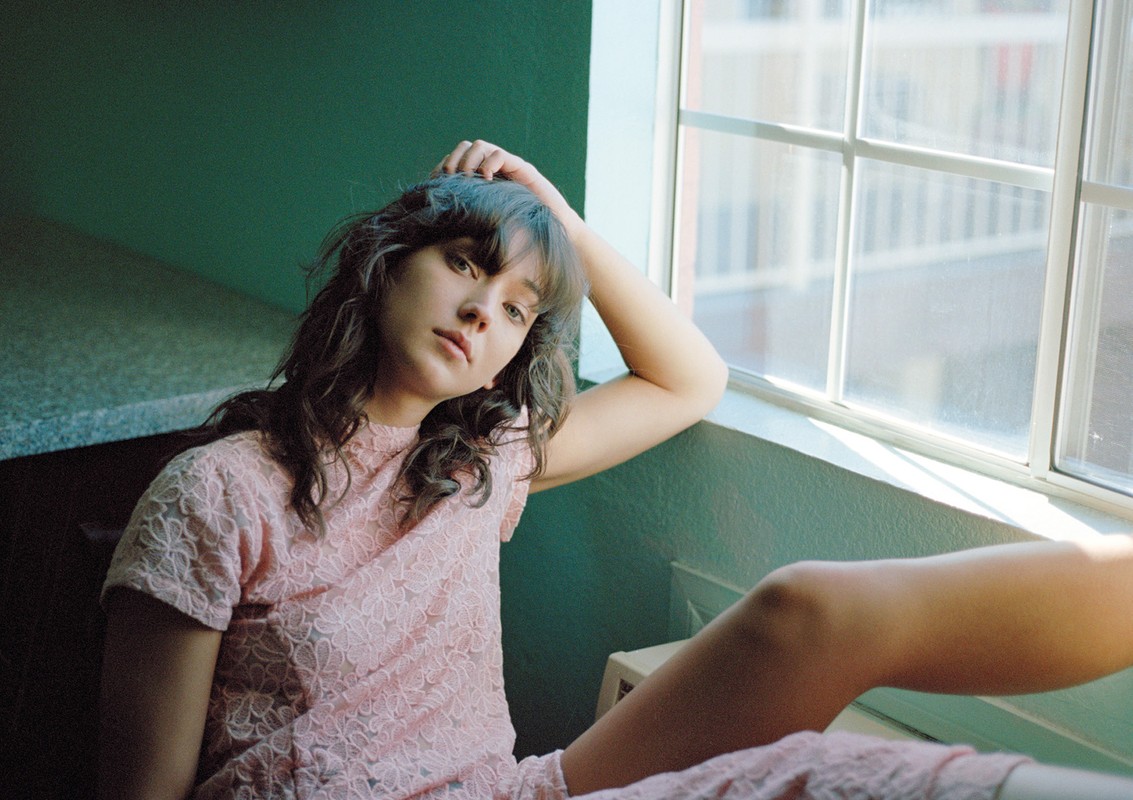 Portrait of a young woman with long wavy brunette hair in a short-sleeve pink lace dress sitting by a window in natural light.