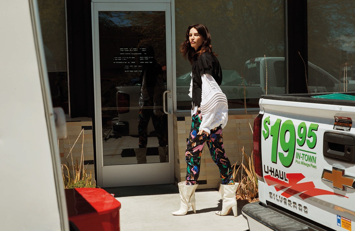 A young woman in patterned pants and white boots walks from a glass door next to a white pickup truck in an outdoor setting.