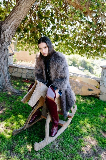 Model in a fur coat sits on a wooden rocking horse outdoors in natural light. Dark hair, burgundy boots, and green grass visible.