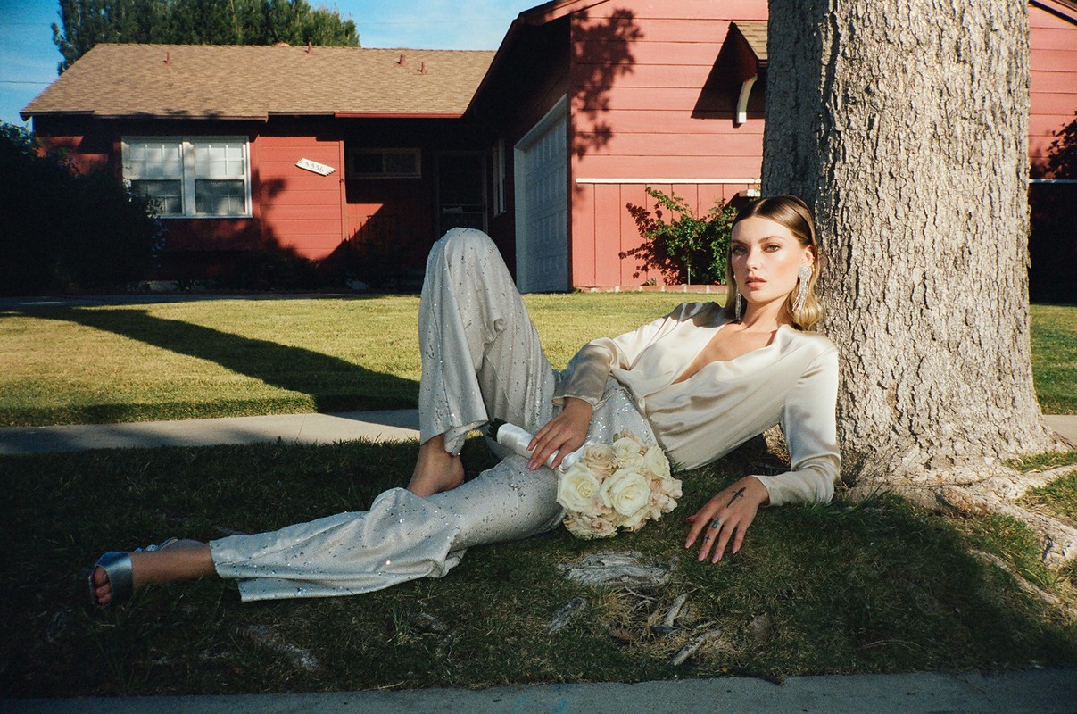 A young adult woman wearing a two-piece pant suit lays on grass beside a tree in front of a red brick house with a white bouquet.
