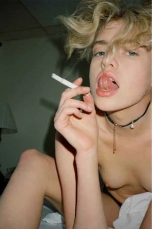 Close-up portrait of a blonde model sticking out their tongue and holding a cigarette with makeup by Karina Moore.